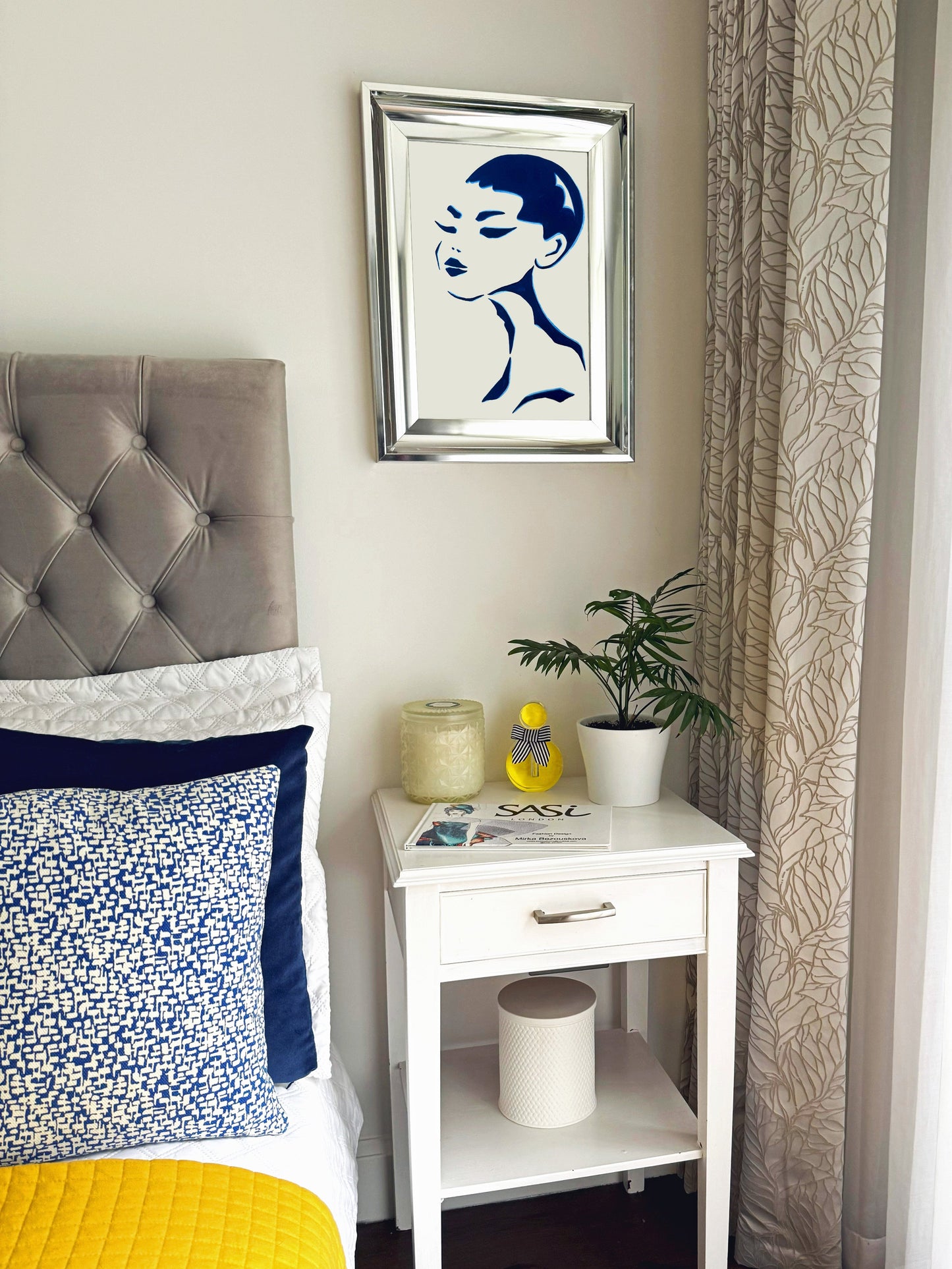 Bedroom with gray upholstered headboard, yellow bedding, white nightstand with decor items, and framed art on wall of a female abstract portrait.