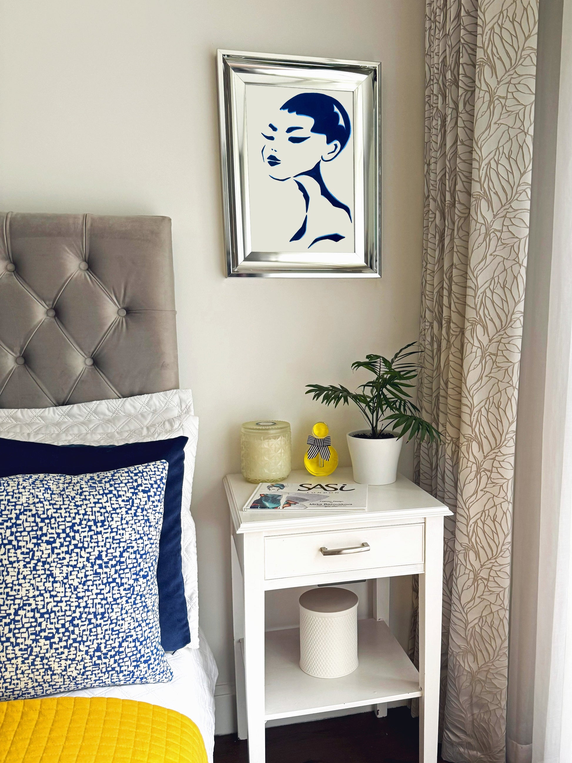 Bedroom with gray upholstered headboard, yellow bedding, white nightstand with decor items, and framed art on wall of a female abstract portrait.