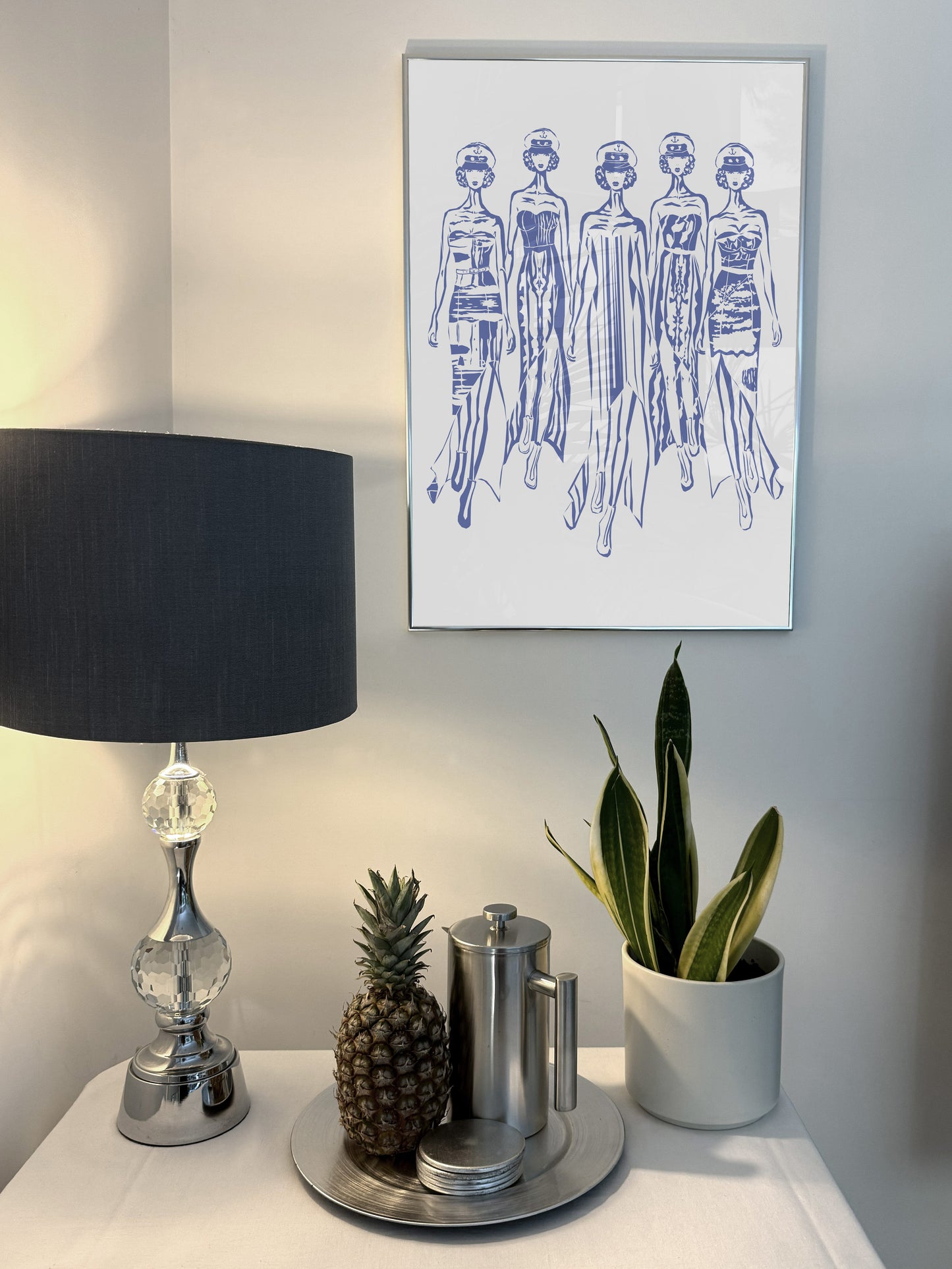 Abstract figurative art, blue acrylic, female catwalk. Decorative table with lamp, plants, and small items against a wall with framed art. 