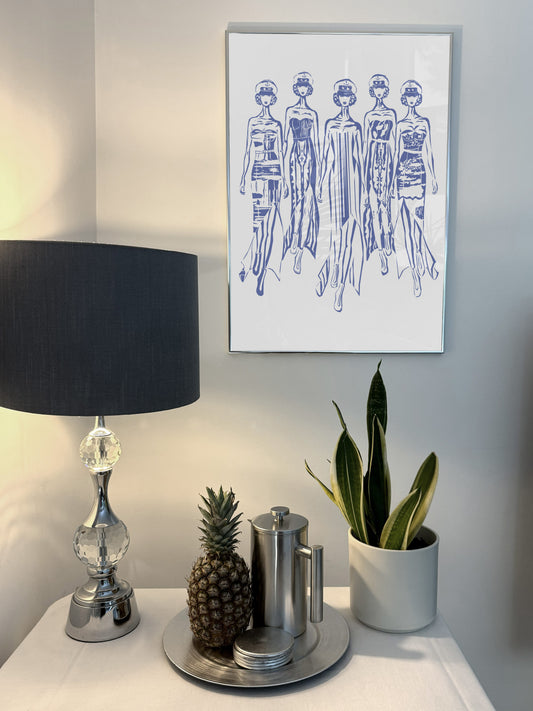Abstract figurative art, blue acrylic, female catwalk. Decorative table with lamp, plants, and small items against a wall with framed art. 