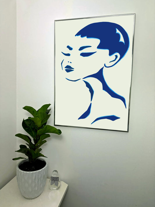 Framed blue and white abstract art of a face on a wall with a plant and a small perfume bottle below.