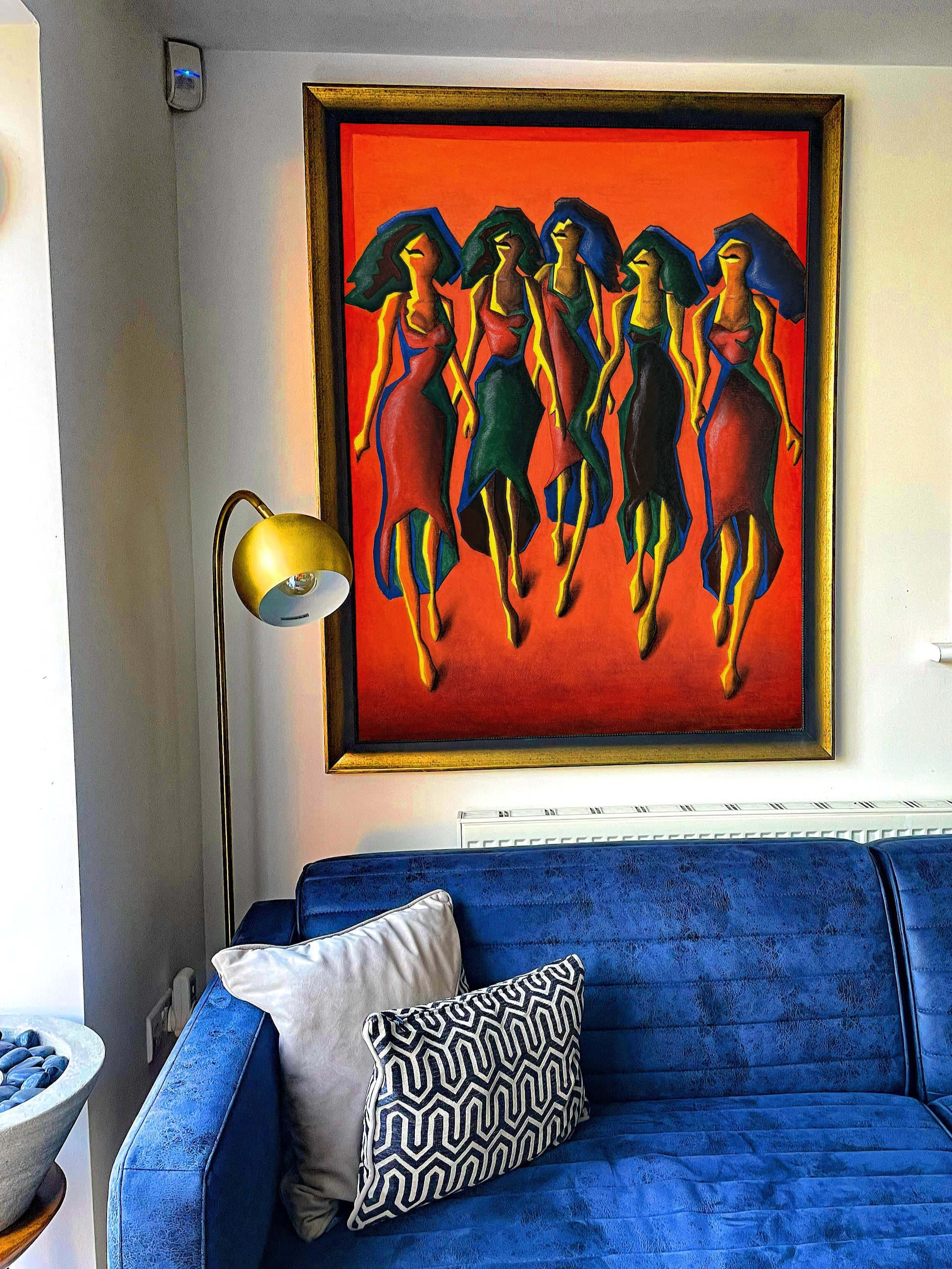 Colorful painting of five women on an orange background above a blue sofa with decorative pillows.