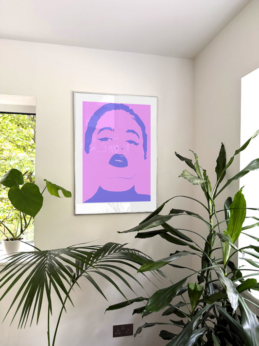 A framed pop-art style poster featuring a stylized face in pink and purple, hanging on a beige wall in a room with green houseplants.