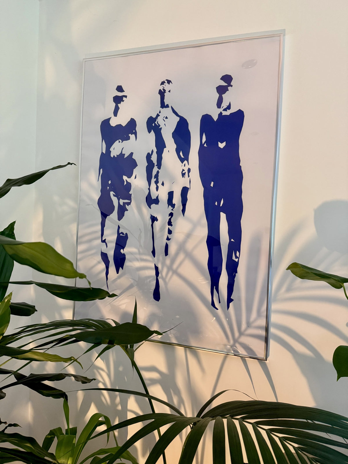 A framed wall art print featuring three abstract blue human silhouettes on a white background, hanging on a light wall with green plant leaves and palm shadows in the foreground. Figurative Art. Walking Men. Abstract Art. Blue Acrylic.